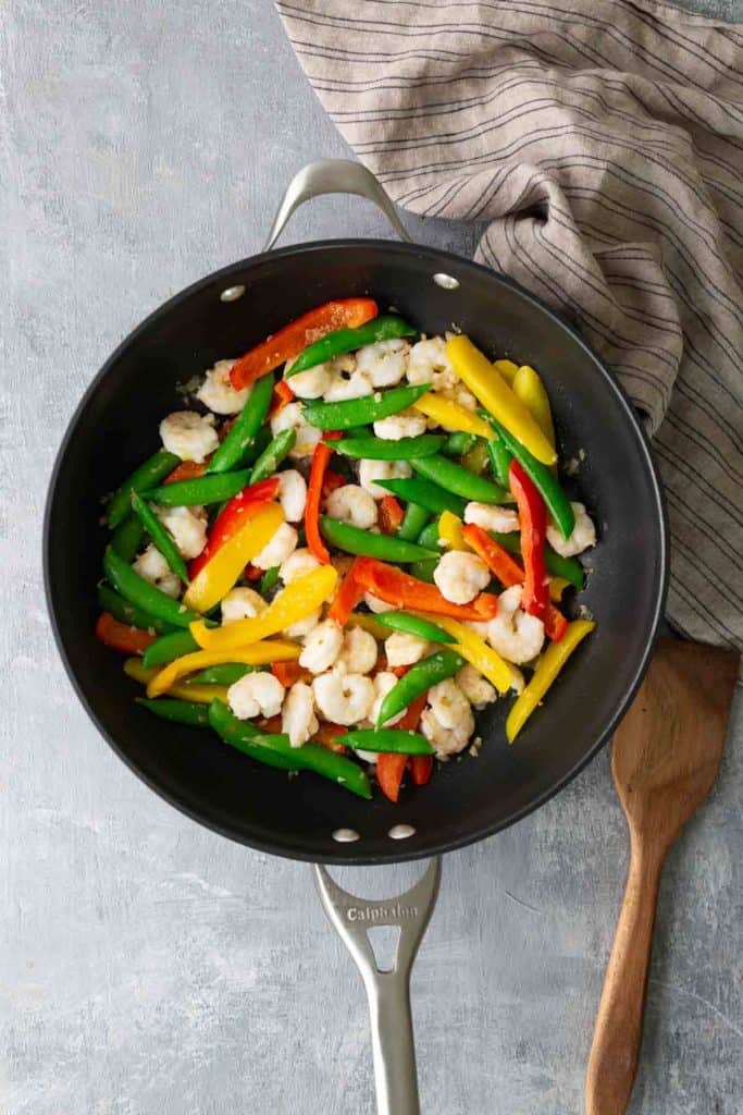 Sauté pan with shrimp, red, yellow, and green bell peppers on a gray surface, next to a striped cloth and a wooden spatula.