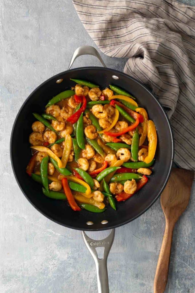 A skillet with cooked shrimp, snap peas, and red, yellow, and green bell pepper strips, placed on a light surface next to a striped cloth and a wooden spatula.
