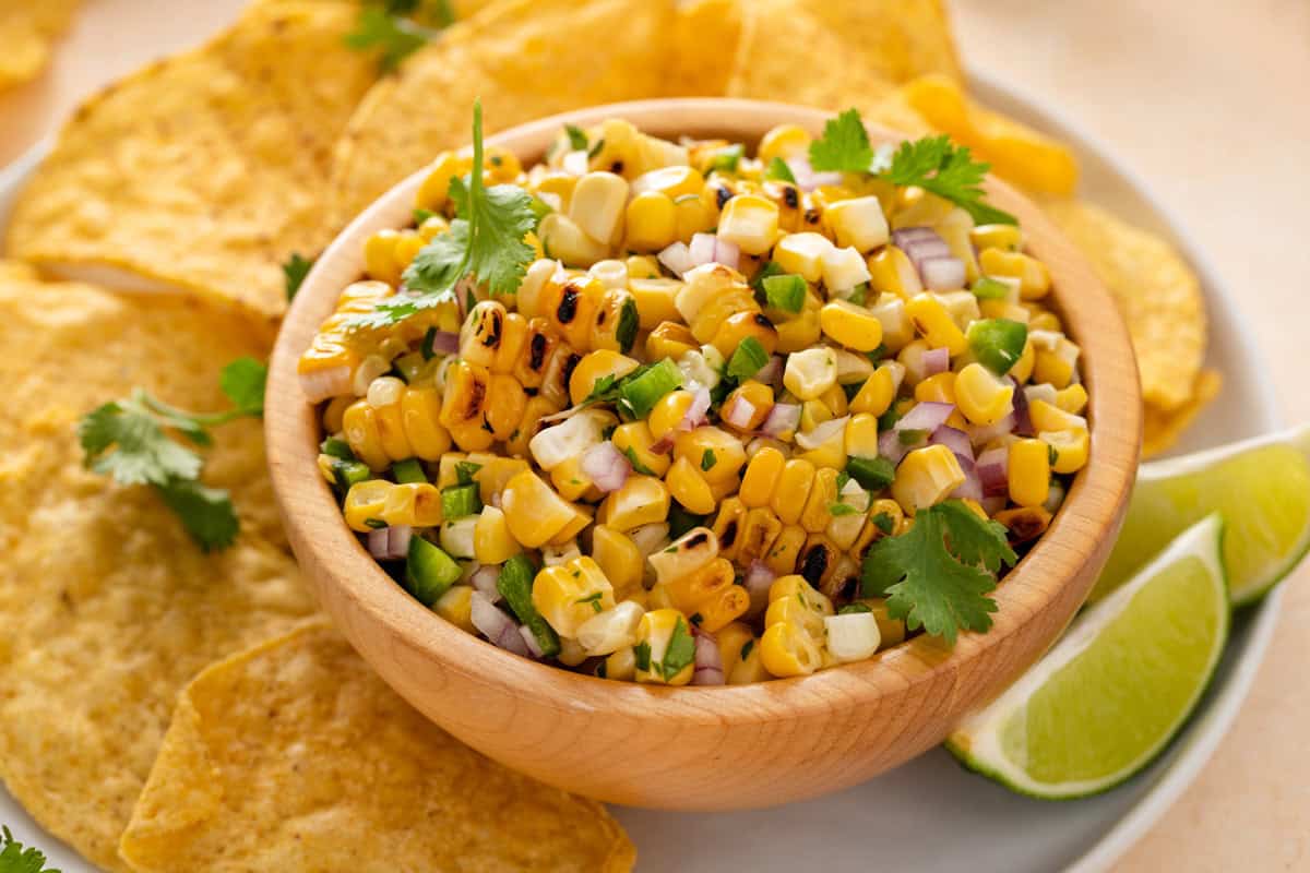 A wooden bowl of grilled corn salsa with chopped onions, jalapeños, and cilantro, surrounded by tortilla chips and lime wedges on a plate.