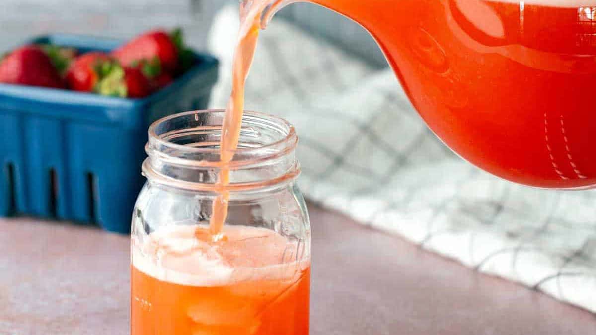 A hand pours bright orange-red juice from a glass pitcher into a mason jar; strawberries and striped straws are nearby.