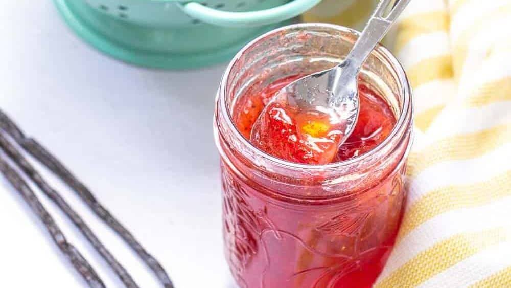 strawberry vanilla jam in a jar with strawberries, vanilla beans and yellow striped kitchen towel on a table