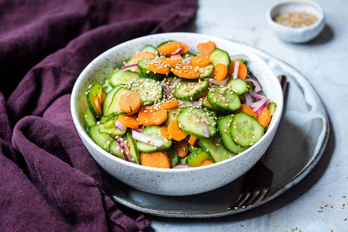A white bowl filled with sliced cucumbers, carrots, and red onions, topped with sesame seeds, placed on a plate with a fork and a dark purple napkin.