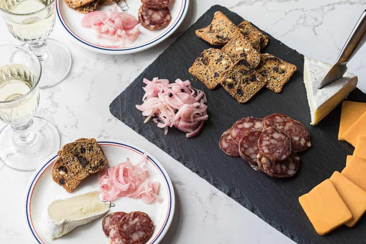 A slate tray with crackers, sliced salami, pickled onions, cheddar, and brie; two plates with similar items and glasses of white wine on a marble table.