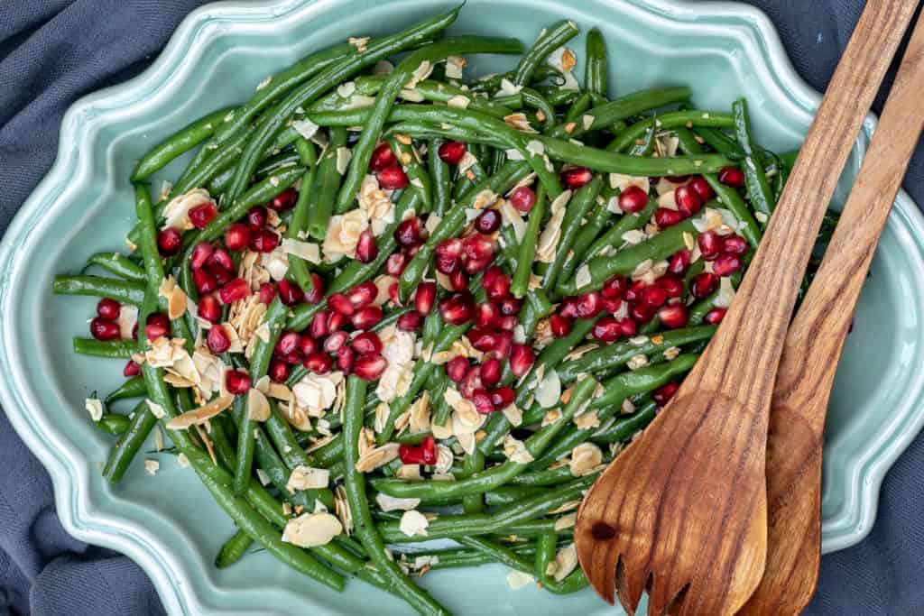 Green bean salad topped with sliced almonds and pomegranate seeds served on a light blue platter with wooden serving utensils.