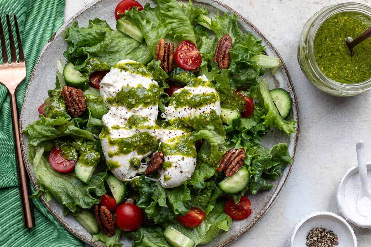 A plate of leafy green salad with cherry tomatoes, cucumber, burrata cheese, candied pecans, and green pesto dressing, next to a jar of pesto and seasoning bowls.
