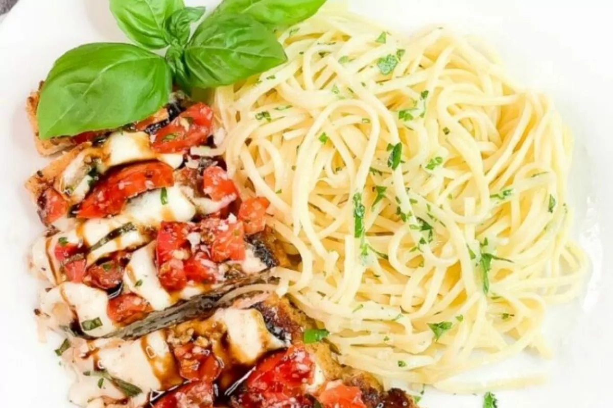 A plate with spaghetti garnished with parsley, grilled chicken topped with tomatoes, mozzarella, balsamic glaze, and basil leaves.