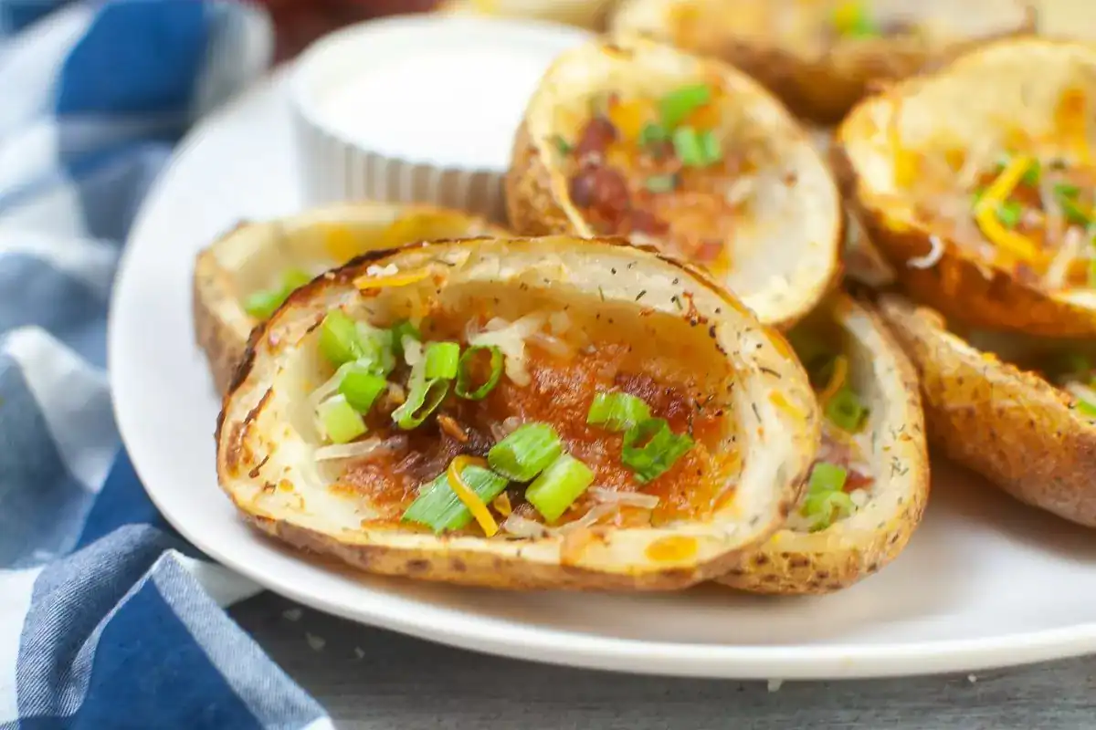 A plate of potato skins topped with melted cheese, bacon bits, and chopped green onions, served with a cup of sour cream.