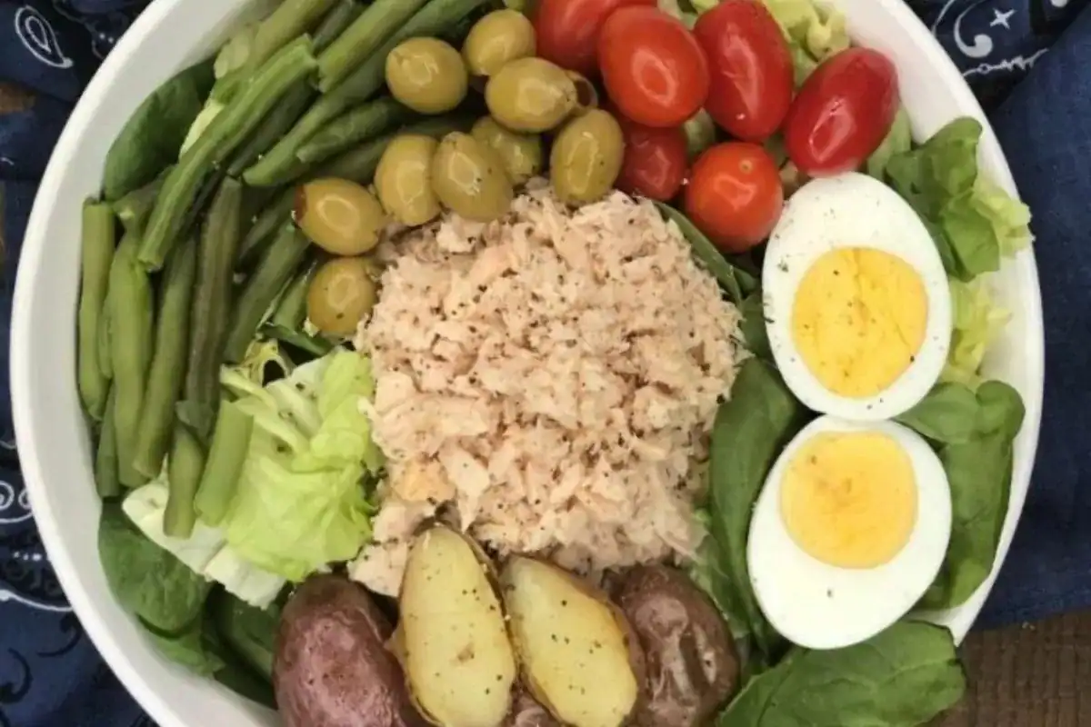 A bowl containing green beans, olives, cherry tomatoes, leafy greens, hard-boiled eggs, boiled potatoes, and flaked tuna.