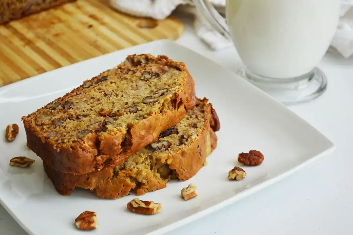 Two slices of banana nut bread on a white plate with pecan pieces, next to a glass mug of milk and a wooden cutting board in the background.