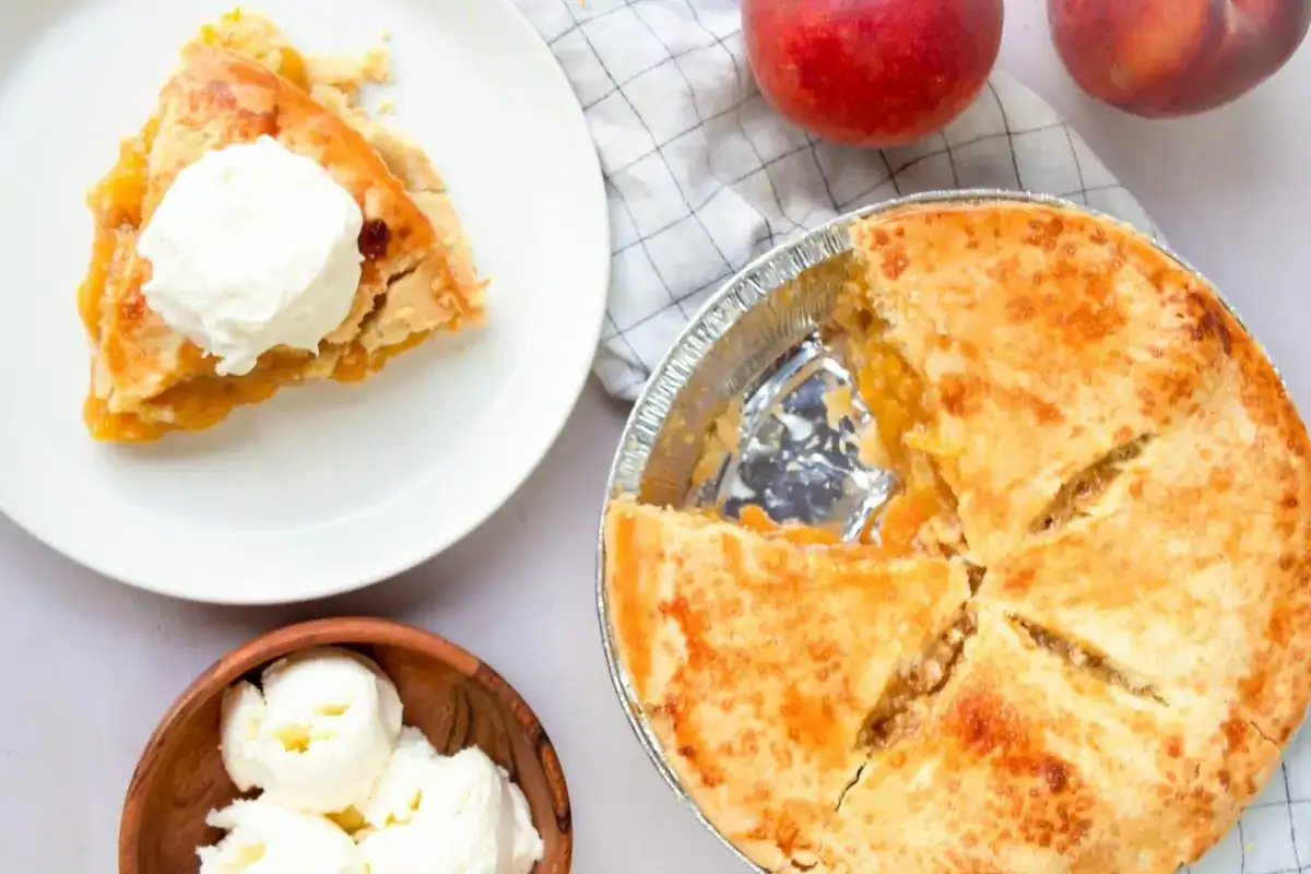 A peach pie with several slices missing sits next to a plate with a slice of pie topped with vanilla ice cream. Nearby are fresh peaches and a bowl of ice cream.