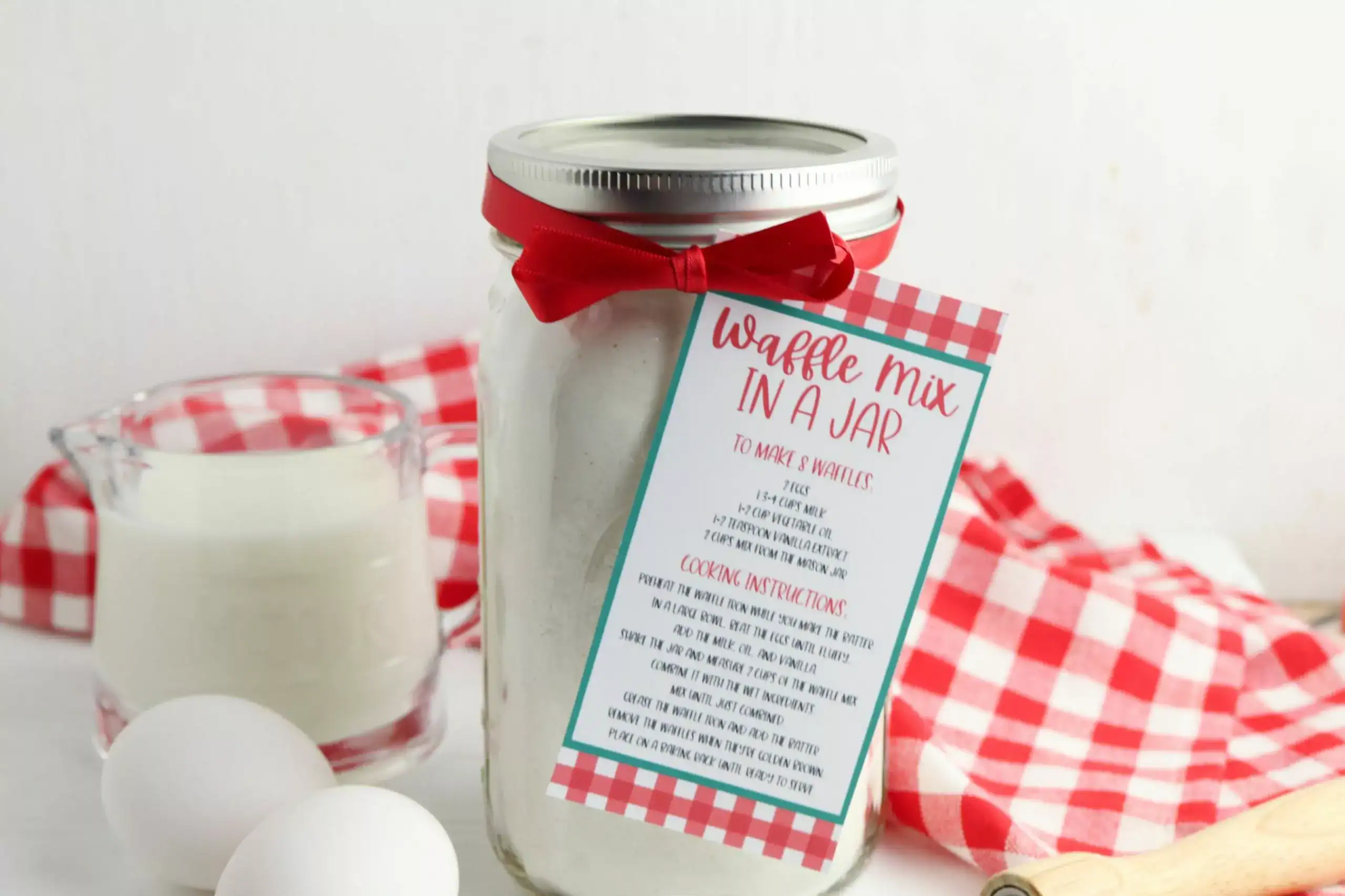 A mason jar labeled "Waffle Mix in a Jar" with a red ribbon, next to eggs, a measuring cup of milk, and a red checkered cloth on a white surface.