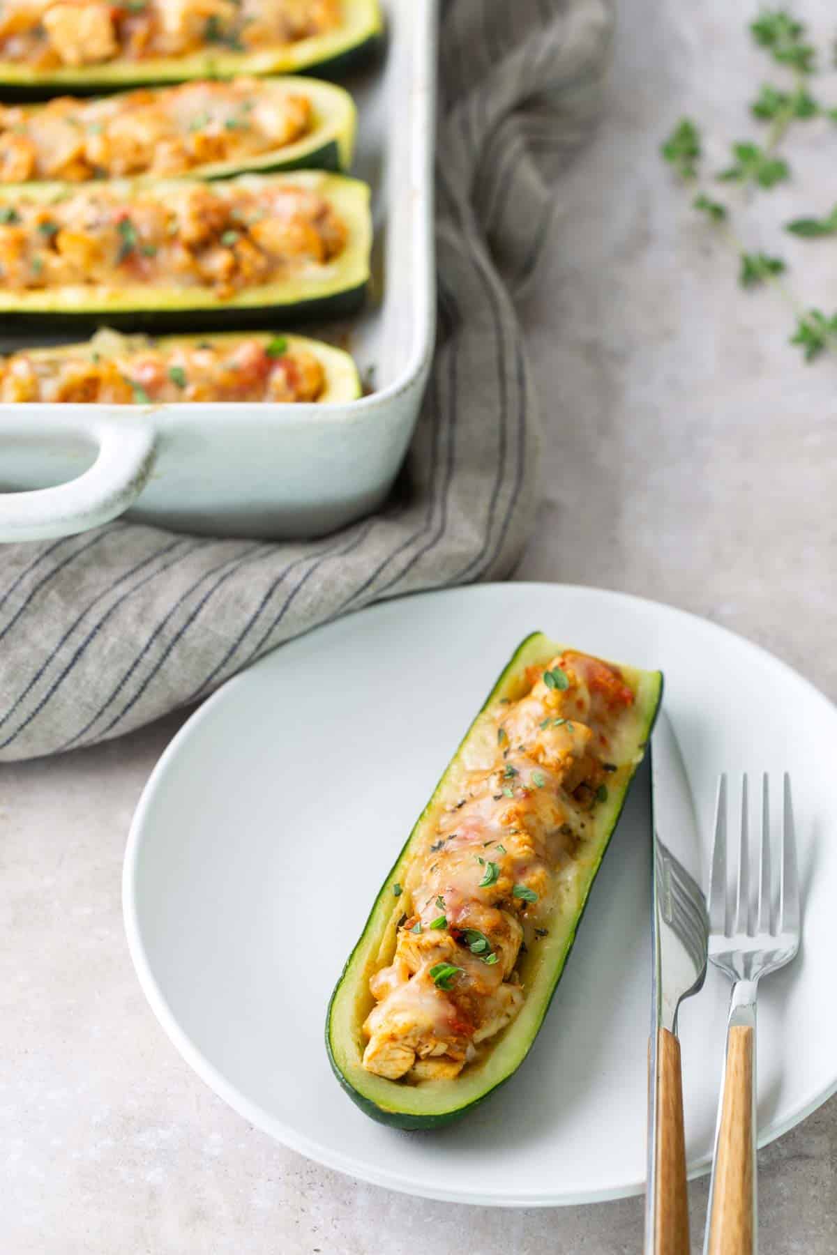 A stuffed zucchini boat filled with a baked, cheesy mixture is served on a white plate with a fork and knife; more stuffed zucchini boats are in a baking dish in the background.