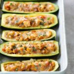 A baking dish with six zucchini boats filled with a seasoned mixture and topped with melted cheese, arranged in two rows.