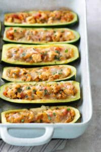 A baking dish with six zucchini boats filled with a seasoned mixture and topped with melted cheese, arranged in two rows.