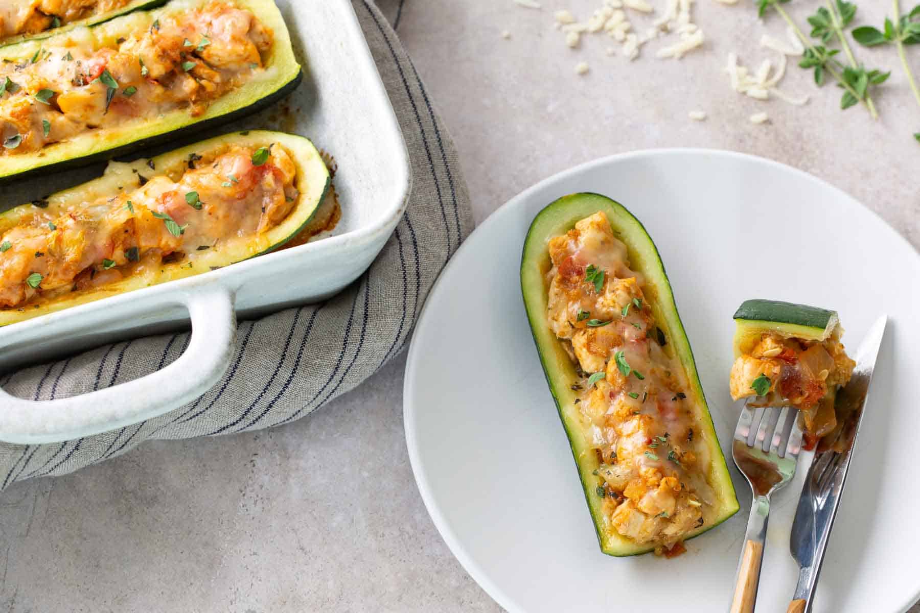 A stuffed zucchini boat filled with a vegetable and cheese mixture is served on a white plate with a fork and knife; more zucchini boats are in a baking dish nearby.