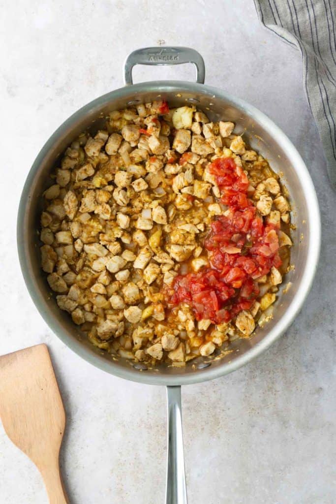 A metal pan filled with cooked diced chicken, chopped onions, and a portion of diced tomatoes on top, sits on a light countertop beside a wooden spatula.