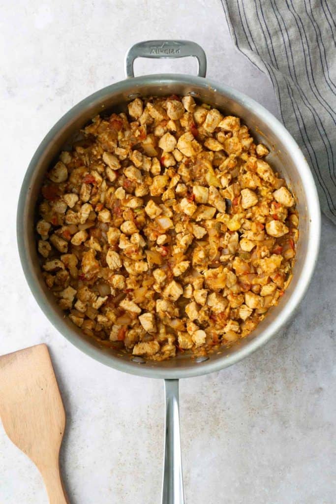 A stainless steel pan filled with cooked diced chicken and vegetables sits on a light surface next to a wooden spatula and a striped cloth.
