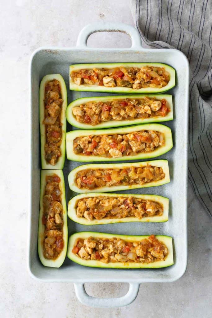 A baking dish with eight zucchini halves, hollowed out and filled with a mixture of diced vegetables and possibly meat, ready to be cooked.