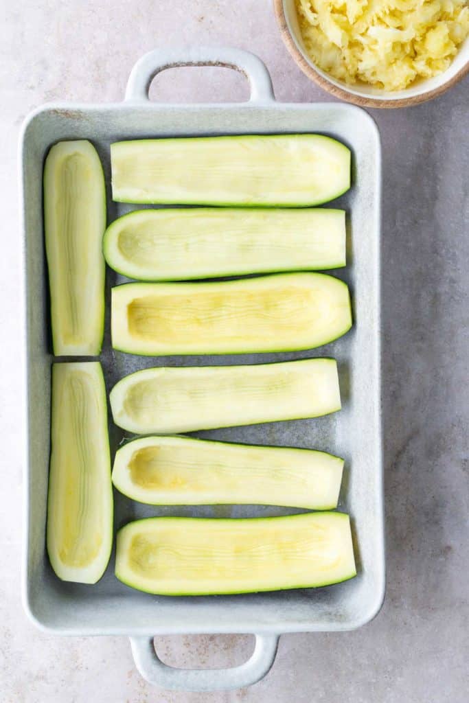 A baking dish with eight hollowed zucchini halves, arranged in two rows; a bowl of scooped zucchini flesh is in the top right corner.
