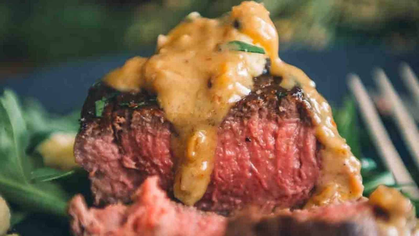 Medium-rare steak topped with a creamy peppercorn sauce, garnished with herbs, on a plate with greens in the background.
