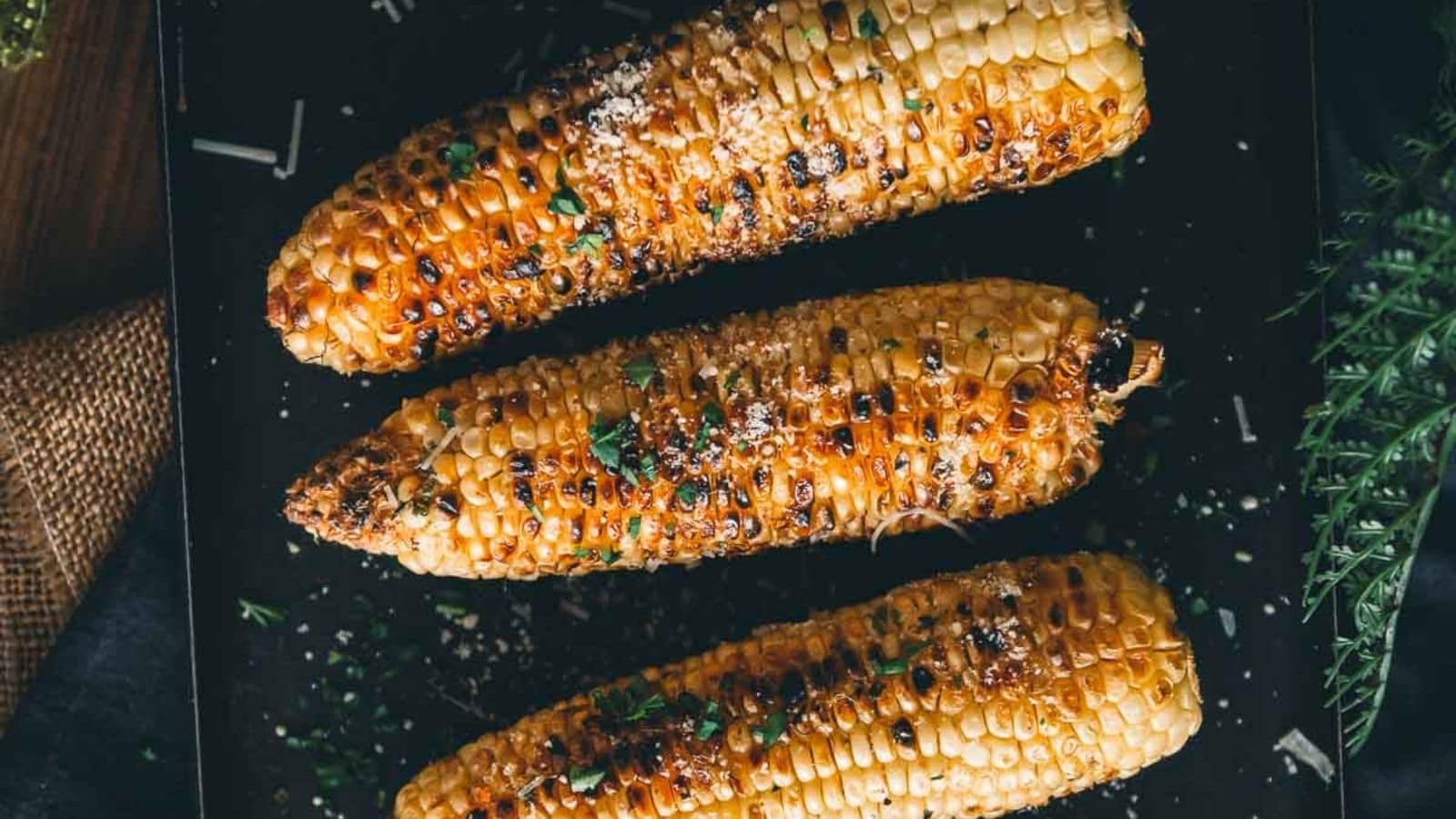 Three grilled corn cobs topped with herbs and grated cheese are arranged on a dark surface.