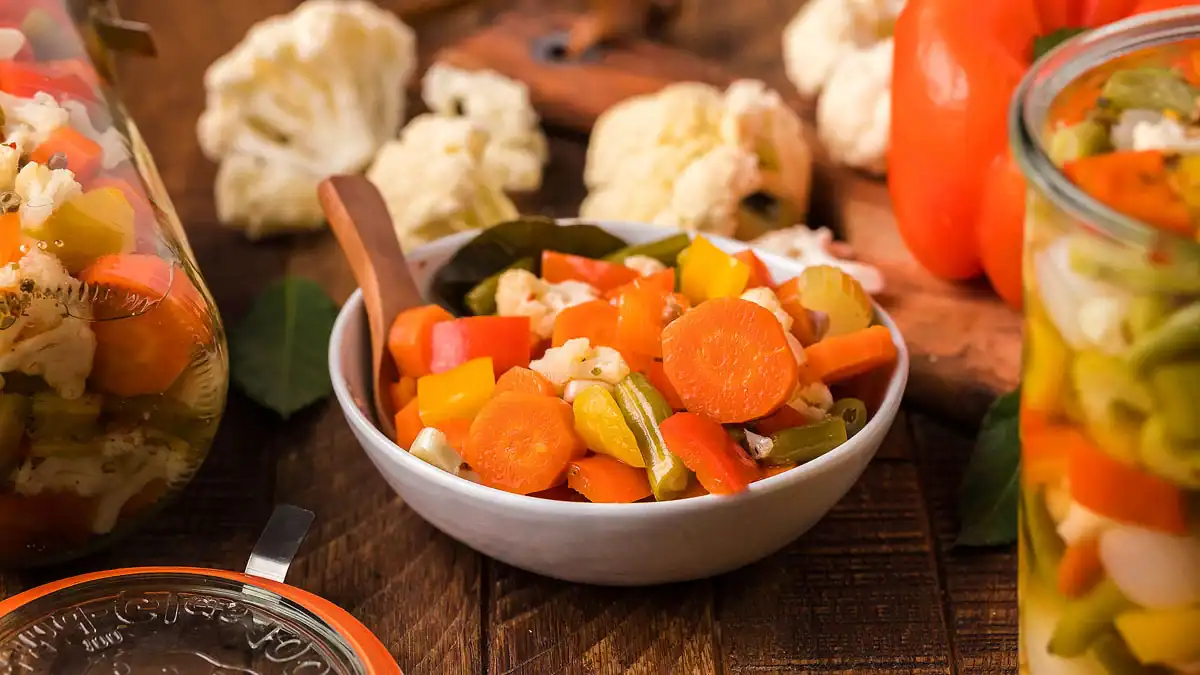 A bowl of mixed pickled vegetables including carrots, cauliflower, celery, and peppers sits on a wooden table with fresh vegetables in the background.