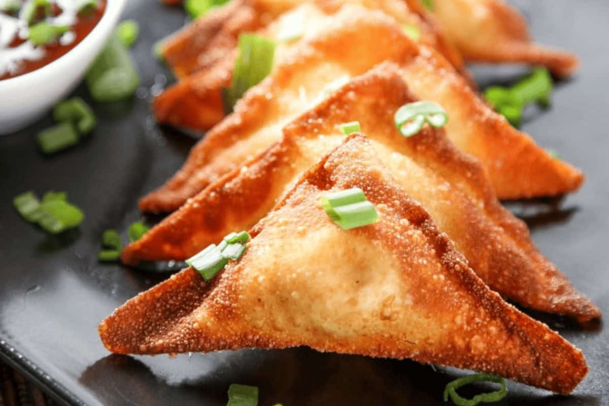 Four golden-brown fried samosas arranged in a row on a black plate, garnished with chopped green onions; a bowl of dipping sauce is partially visible in the corner.