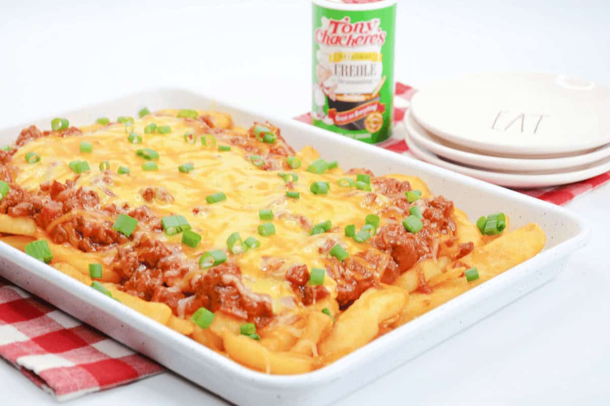 A tray of chili cheese fries topped with chopped green onions sits on a red checkered cloth, with seasoning and plates in the background.