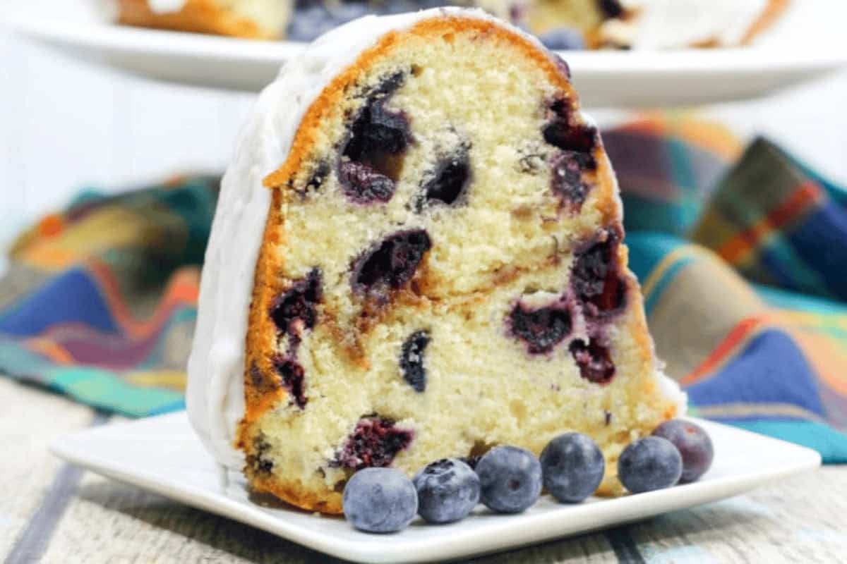 A slice of blueberry bundt cake with white icing on a square plate, garnished with fresh blueberries, with a colorful cloth in the background.