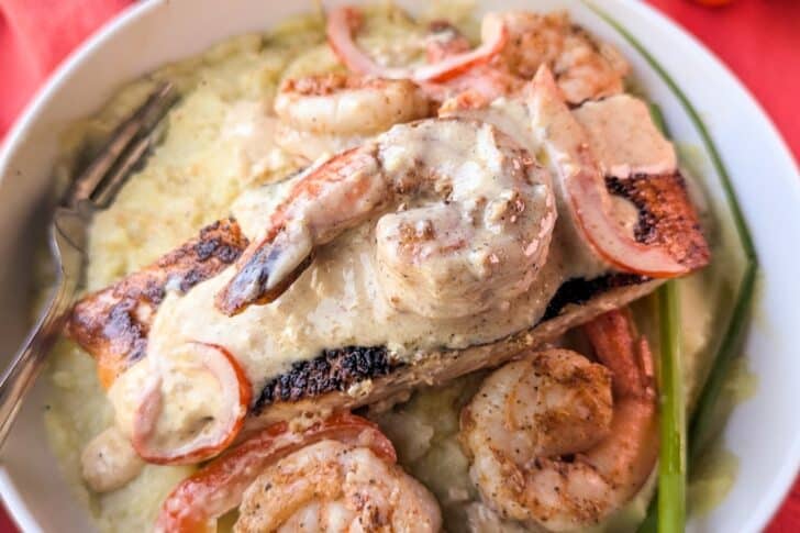 A bowl of grilled shrimp and salmon topped with creamy sauce, served over mashed potatoes with green beans and red pepper slices.