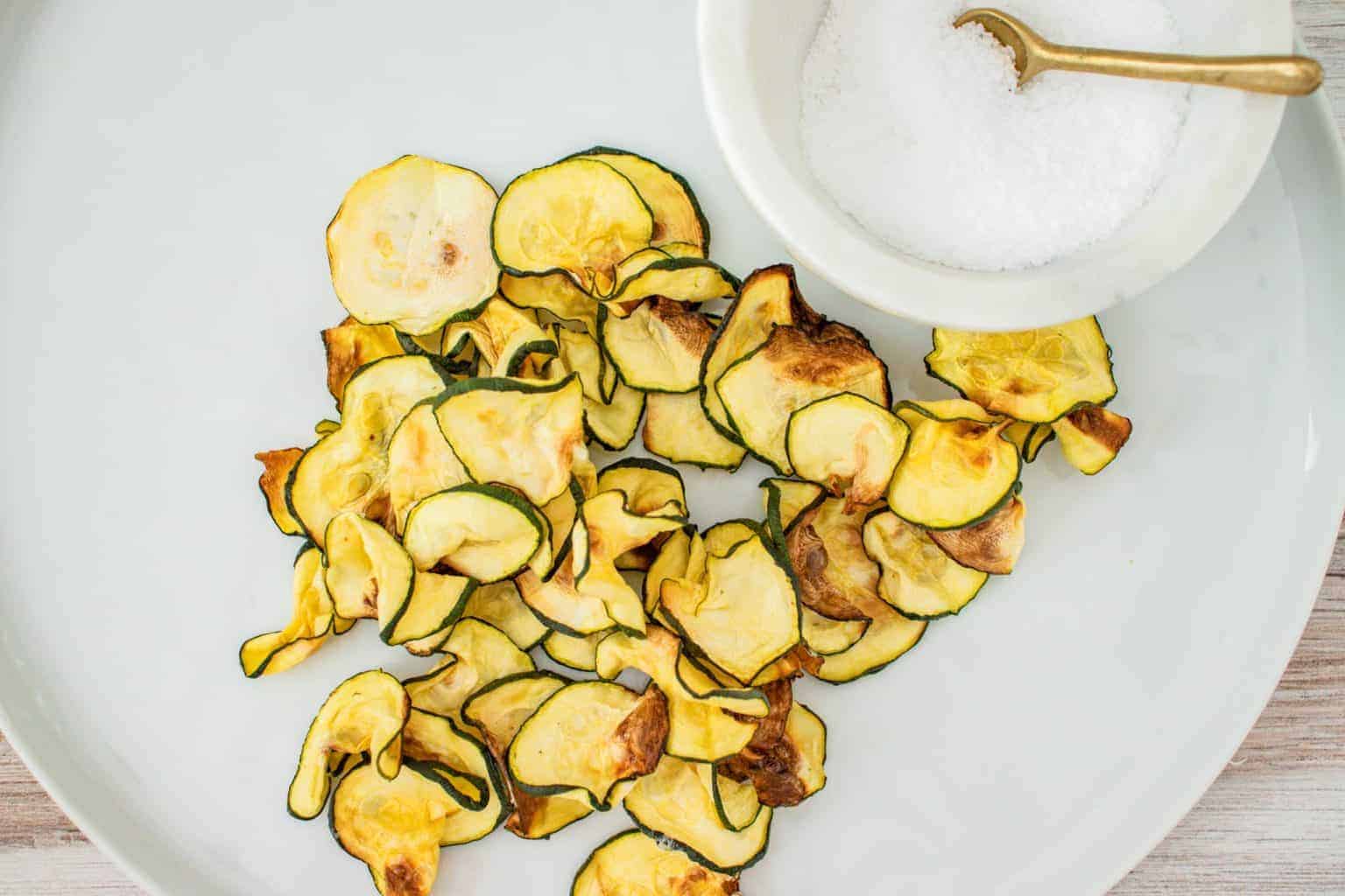 A pile of baked zucchini chips is arranged on a white plate next to a small bowl of salt with a gold spoon.