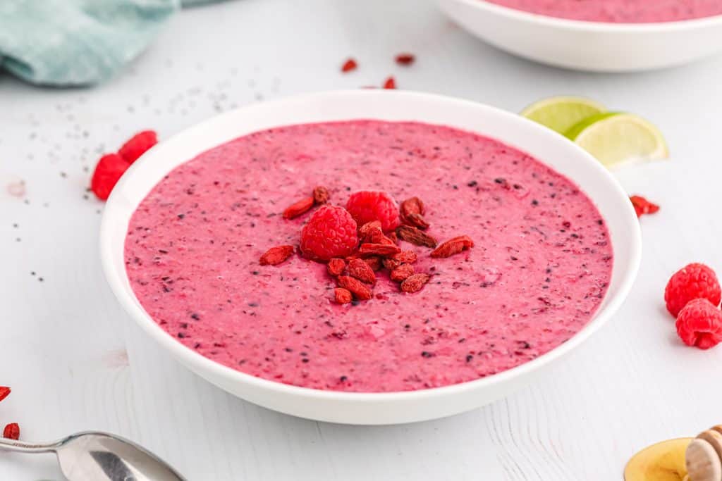 A white bowl filled with a thick pink smoothie topped with raspberries and goji berries sits on a white surface with a spoon and fruit nearby.