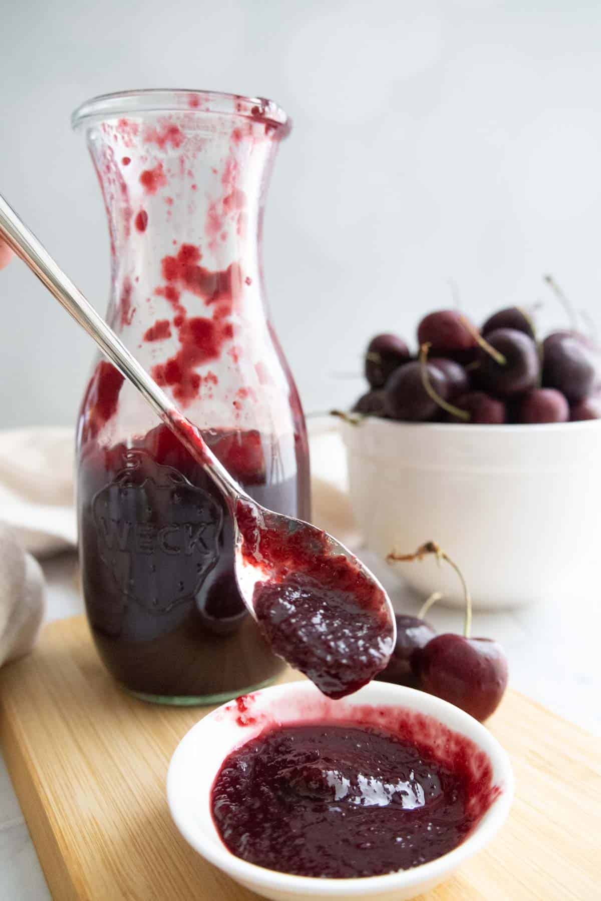 A glass jar of cherry sauce sits on a wooden board, with some sauce on a spoon held over a small dish; a bowl of fresh cherries is in the background.