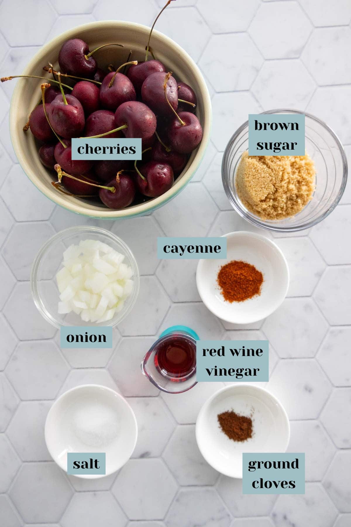 A bowl of cherries, and small bowls containing brown sugar, diced onion, cayenne, red wine vinegar, salt, and ground cloves, arranged on a white hexagon tile surface.