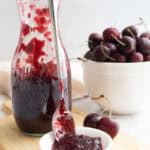 A glass jar of cherry jam with a spoon rests beside a small dish of jam; a white bowl of fresh cherries is in the background on a wooden board.