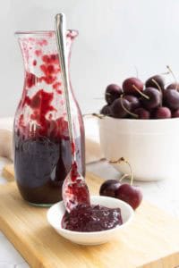 A glass jar of cherry jam with a spoon rests beside a small dish of jam; a white bowl of fresh cherries is in the background on a wooden board.