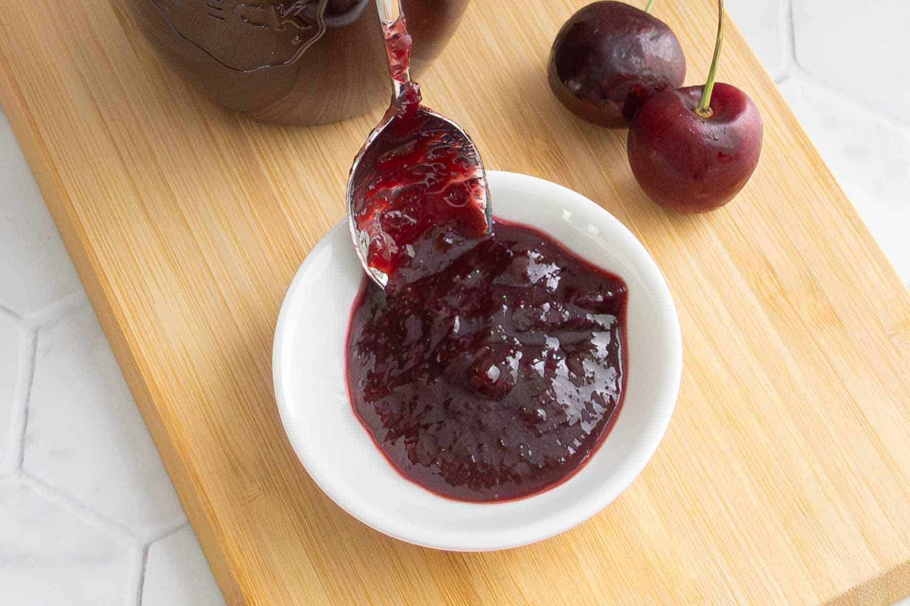 A spoon with cherry jam rests on a small white dish, with fresh cherries and a wooden board in the background.