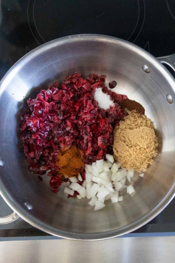 A stainless steel pot on a stove containing chopped onions, dried cranberries, brown sugar, salt, and spices, ready to be mixed and cooked.