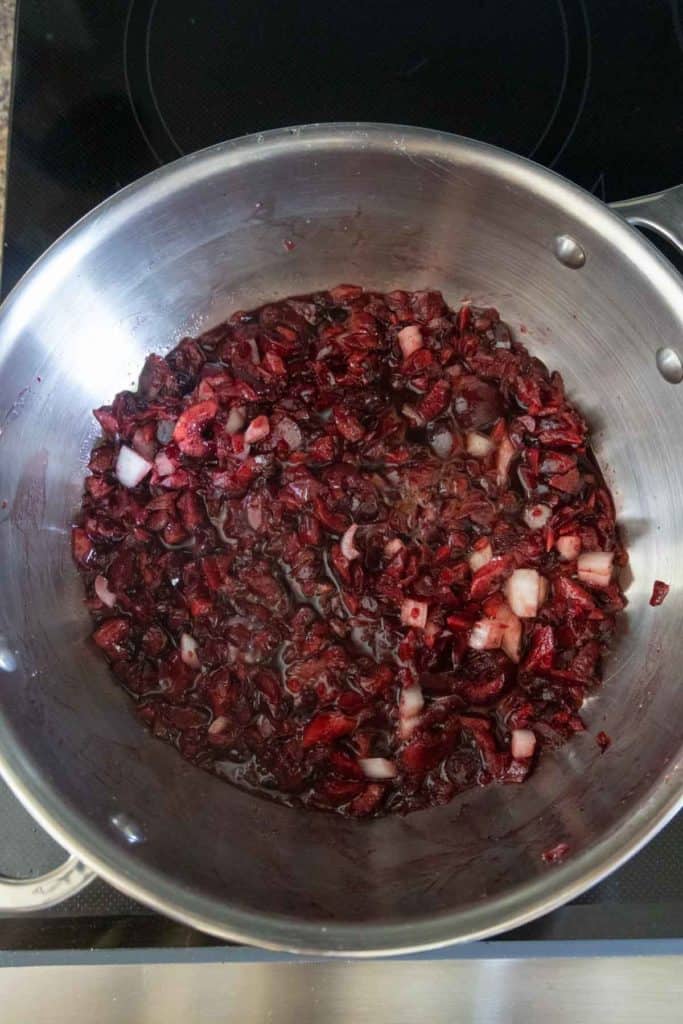 A stainless steel pan containing chopped onions and cranberries being cooked together on a stovetop.