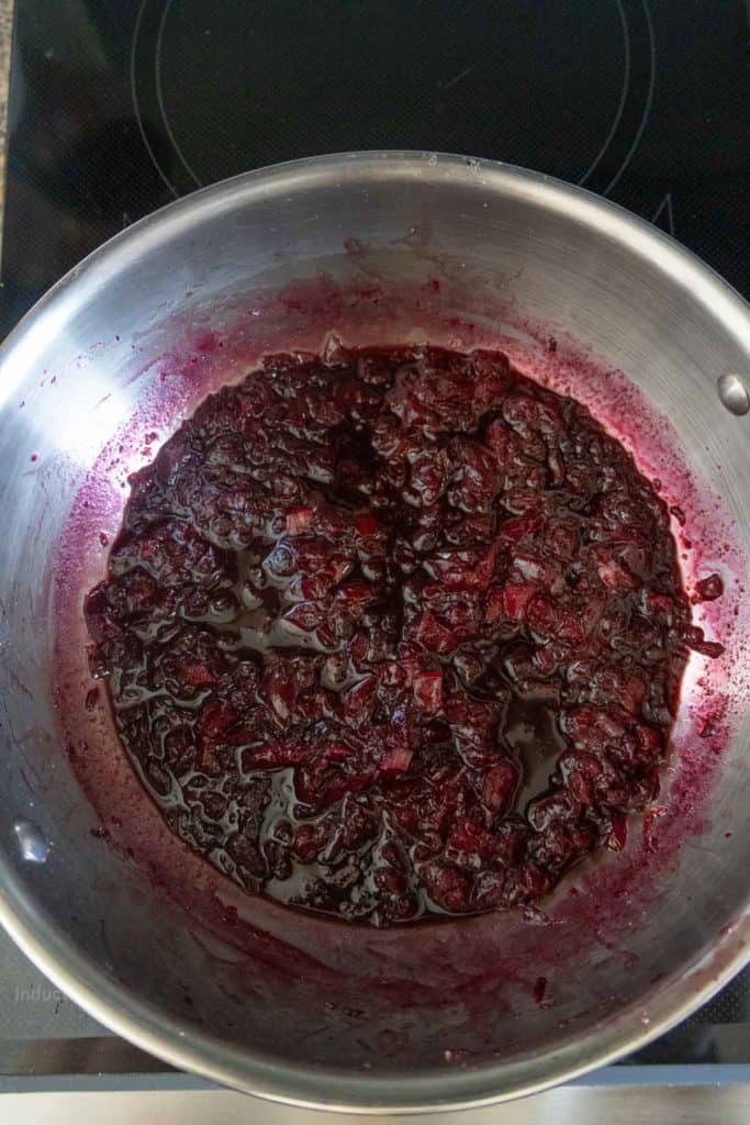 A stainless steel pot on a stovetop contains a dark red, chunky fruit mixture simmmering in liquid.