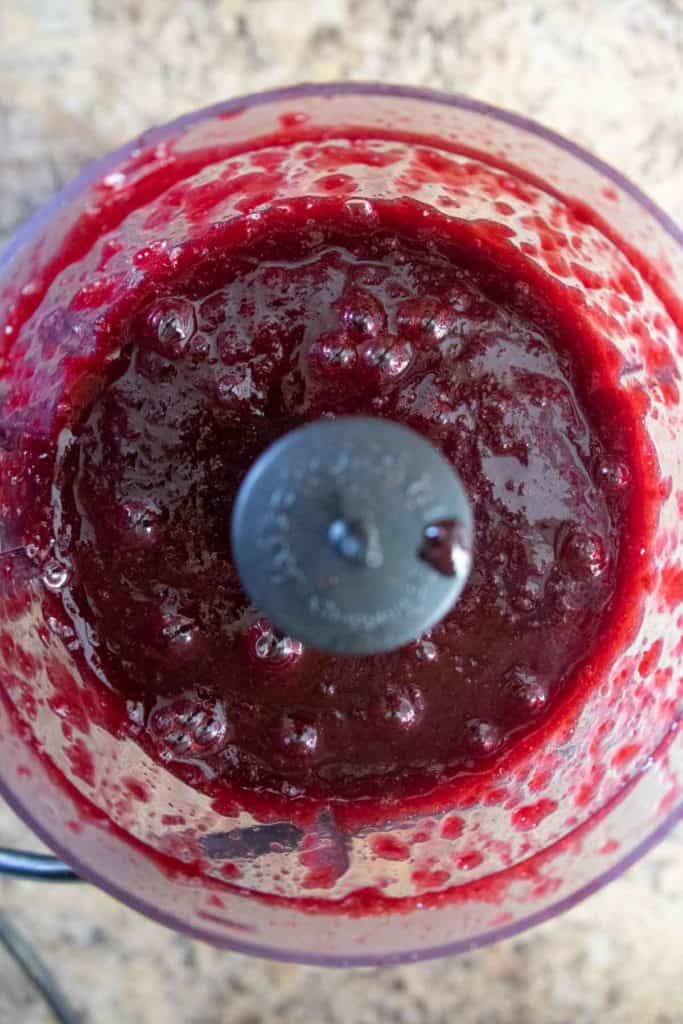A food processor filled with blended red berry sauce, viewed from above on a beige countertop.