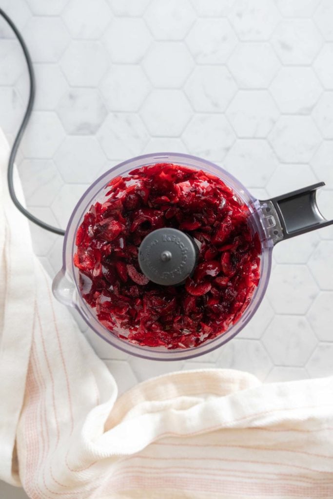 Chopped cranberries in a food processor bowl on a white hexagonal tile countertop beside a striped dish towel.