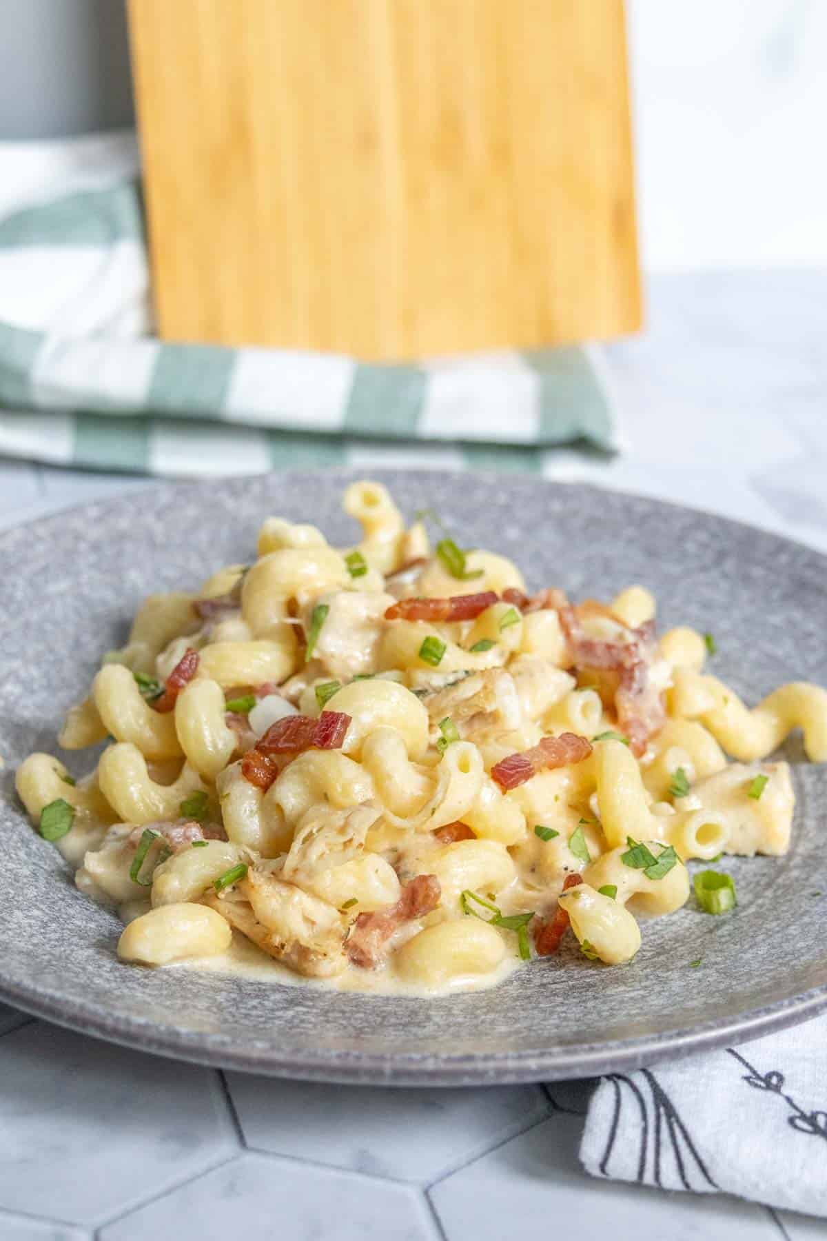 A plate of creamy macaroni pasta mixed with pieces of bacon and garnished with chopped green onions, served on a gray plate.