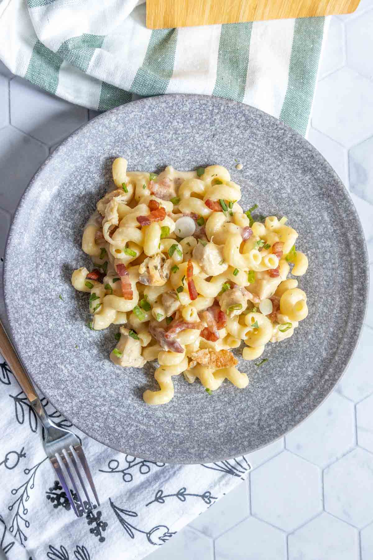 A plate of cavatappi pasta mixed with pieces of chicken, bacon bits, chopped green onions, and a creamy sauce is displayed on a grey plate.