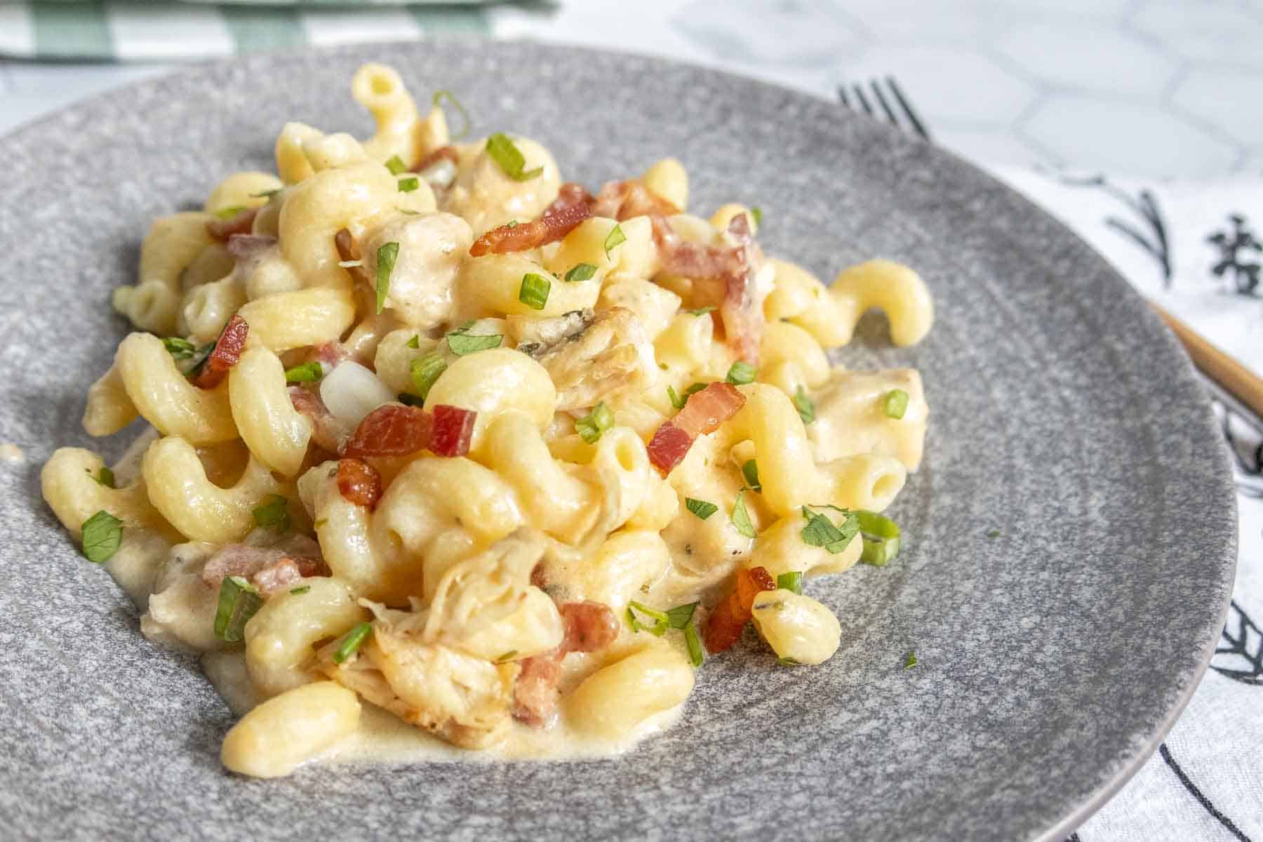 A plate of creamy macaroni pasta with pieces of bacon, shredded chicken, and chopped green herbs on top.
