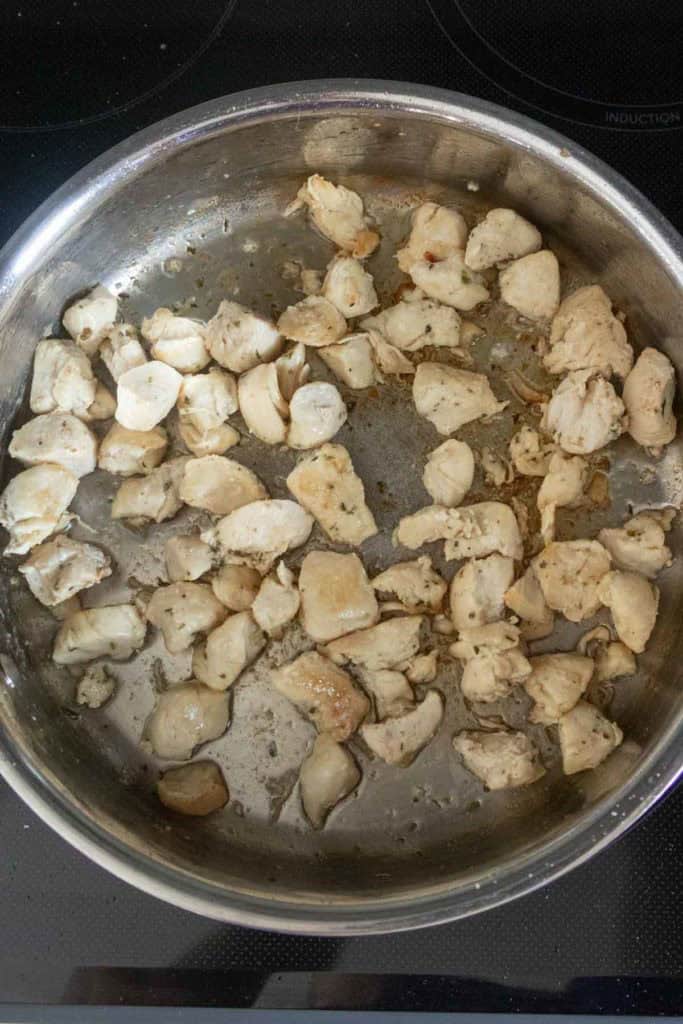 Cubed pieces of chicken are being cooked in a stainless steel skillet on a stovetop.