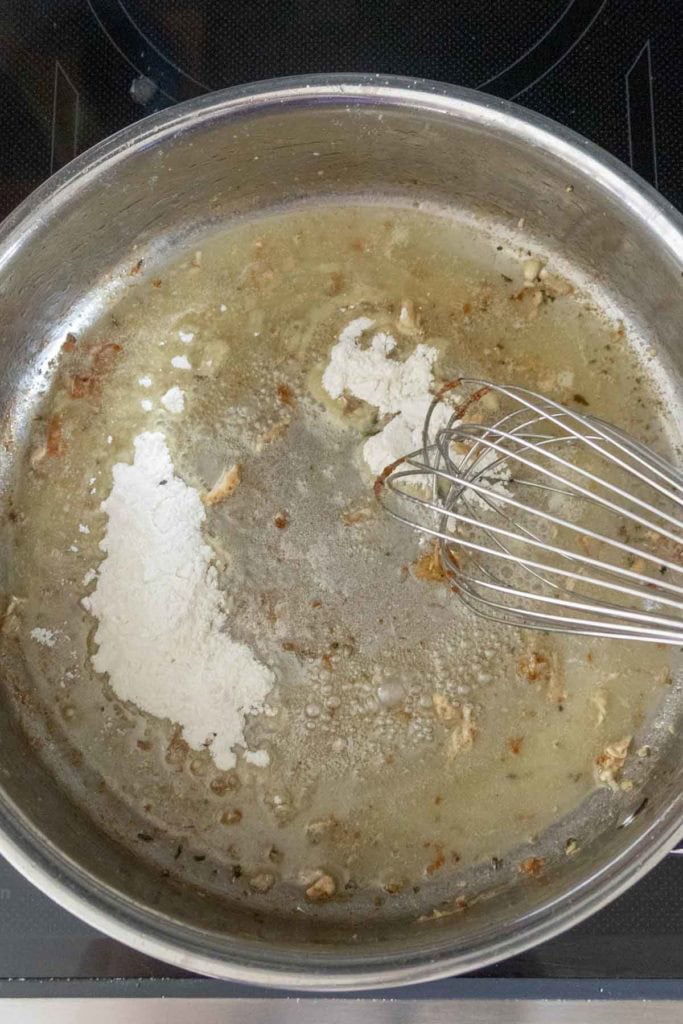 A metal whisk stirs flour into melted butter and browned bits in a stainless steel pan on a stovetop.