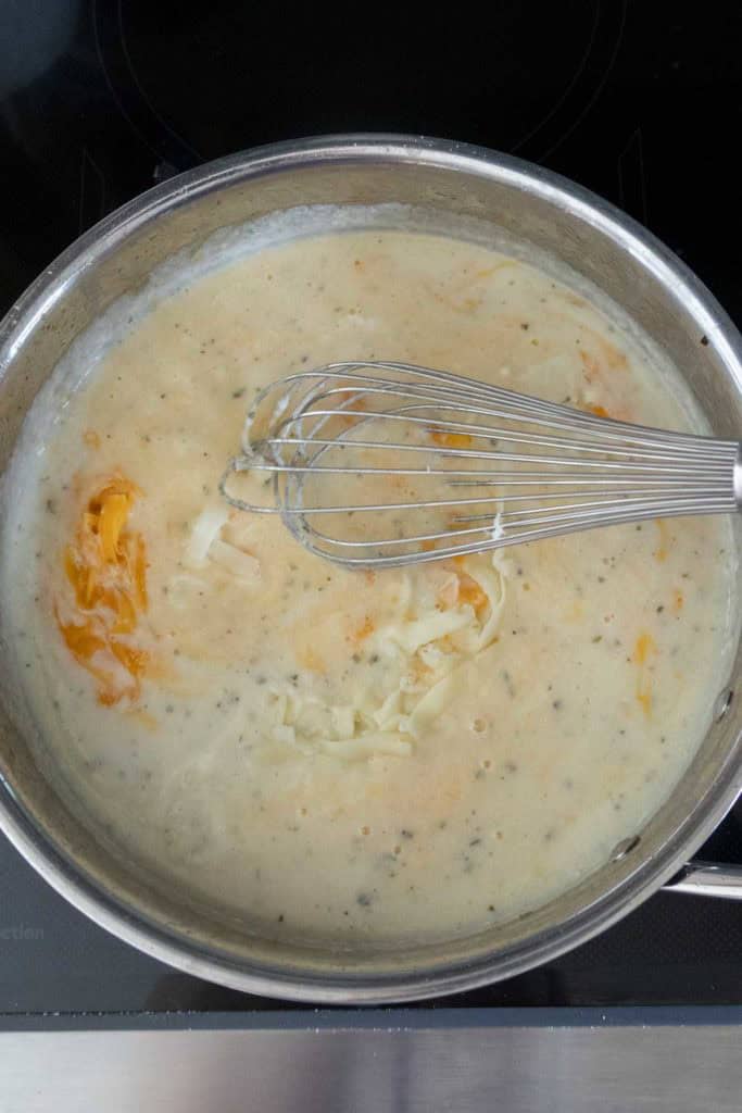 A metal whisk stirring shredded cheese into a creamy sauce in a stainless steel pan on a stovetop.
