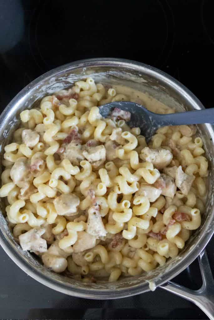 A pot of cooked cavatappi pasta mixed with creamy sauce, diced chicken, and bits of bacon, with a black spoon resting inside.