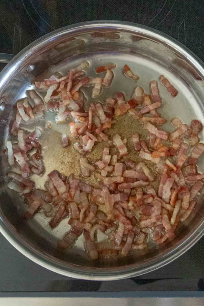 Strips of bacon are being cooked and browned in a stainless steel pan on a stovetop.